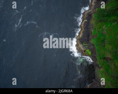 Aus der Luft geschossen. Dunkelblaue Meeresoberfläche mit hellen Wellen und weißen Lichtwellen. Ruhig. Raue hügelige Küste mit grünem Gras überwuchert. Wunderschönes Nat Stockfoto