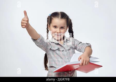 Das kleine Mädchen hält ein rotes Buch in den Händen und gibt einen Daumen nach oben. Stockfoto