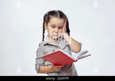 Das kleine schwarzhaarige Mädchen hält ein rotes Buch und legt ihre Hand an den Kopf. Stockfoto