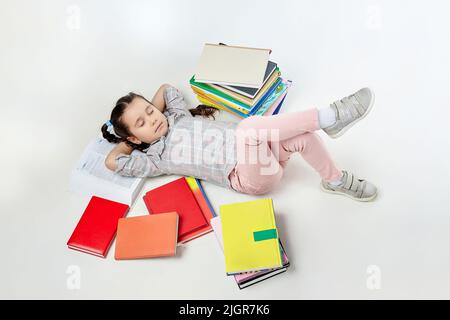 Charmantes kleines Mädchen schläft in einem Studio auf weißem Hintergrund, viele Bücher sind um sie verstreut. Stockfoto