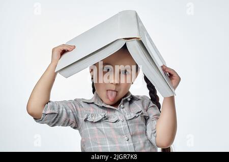 Lächelndes kleines Mädchen mit geflochtenen Zöpfen hält ein großes Buch über ihren Kopf und zeigt ihre Zunge. Stockfoto