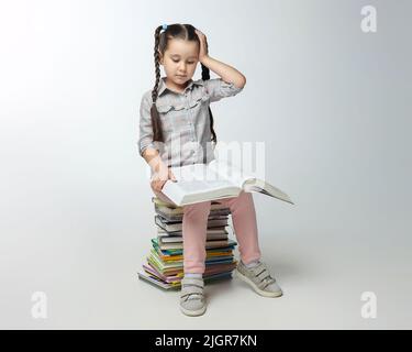 Das kleine Mädchen mit geflochtenen Zöpfen sitzt auf einem großen Stapel von Büchern und drückt ihre Hand an den Kopf. Stockfoto