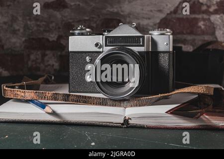 Auf einem abgenutzten und zerkratzten Tisch befindet sich eine alte und schmutzige Analogkamera auf einem offenen Notizbuch, auf dem sich auch ein Bleistift und fotografische Negative befinden Stockfoto