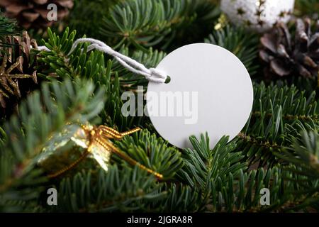 Weihnachtliche runde weiße Tag-Mockup mit weißer Kordel, Nahaufnahme auf natürlichem Tannenzweig, mit Zapfen und Weihnachtsdekoration, Weihnachtsverkauf-Konzept. Bl Stockfoto