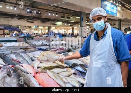 Porträt eines hübschen jungen erwachsenen Fischers aus dem Nahen Osten, der eine Maske trägt, die Kamera anschaut und frischen Fisch auf dem traditionellen Fischmarkt verkauft. VAE Stockfoto