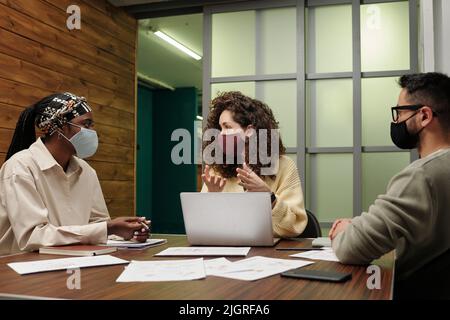 Gruppe von jungen interrassischen Ökonomen in der Casualwear und schützende Gesichtsmasken, die Diskussion über neue Ideen während der Arbeit über Projekt Stockfoto