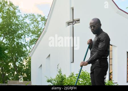 Junger ernsthafter afroamerikanischer Pastor in schwarzen Hosen und Hemden und Brillen, der im Garten beim Kirchengebäude mit Rechen arbeitet Stockfoto