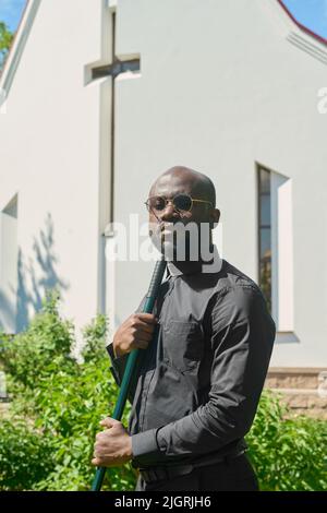 Junger ernsthafter Pastor afroamerikanischer Ethnie hält Arbeitswerkzeug, während er im Kirchengarten gegen weißes Gebäude steht und auf die Kamera schaut Stockfoto