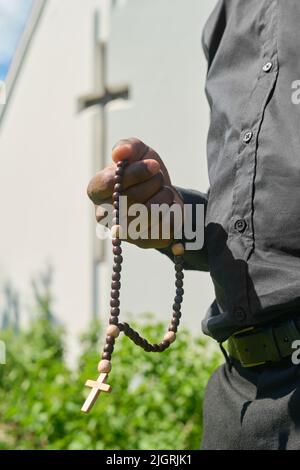 Hand eines jungen afroamerikanischen Priesters mit hölzernen Rosenkranzperlen, die im Garten neben dem Kirchengebäude Gebet sagen oder stilles Gebet üben Stockfoto