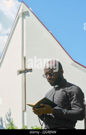 Ernsthafter afroamerikanischer Mann mit offener Bibel und hölzernen Rosenkranz-Perlen, der Verse aus dem Evangelium gegen den Bau der Kirche liest Stockfoto