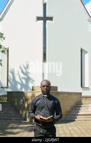 Junger ernsthafter Priester mit offener Bibelevangelisierung oder erzählender Predigt, während er vor dem Gebäude der katholischen Kirche steht Stockfoto