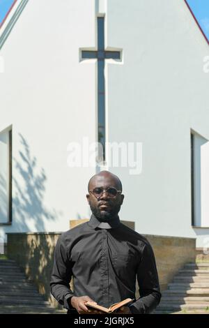 Ernsthafter schwarzer Mann mit offenem Evangelium in den Händen und Blick auf die Kamera gegen das weiße Gebäude der lutherischen Kirche mit Kreuz auf dem vorderen Teil Stockfoto