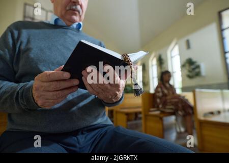 Hände eines älteren männlichen Pfarrers der katholischen Kirche, der die Heilige Bibel in einem schwarzen Ledereinband mit hölzernen Rosenkranzperlen zwischen den Seiten aufhält Stockfoto