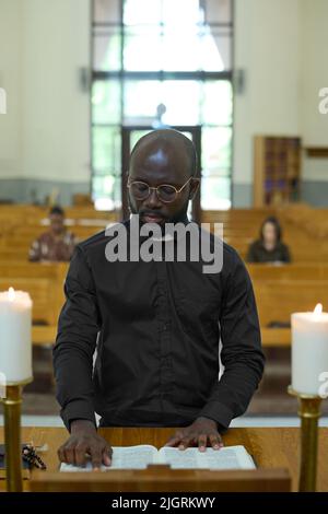 Junger afroamerikanischer Mann in schwarzem Hemd mit Bürokragenkragen, der während der Liturgie eines der vier Evangelien oder eines anderen Buches aus der Heiligen Bibel liest Stockfoto