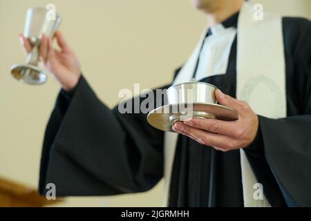 Katholischer Priester, der Becher mit Wein und ungesäuertem Brot hält, das Fleisch und Blut des Erlösers symbolisiert und für die Gemeinschaft der Gemeindemitglieder vorbereitet ist Stockfoto