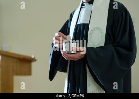 Priester der katholischen Kirche in Soutane hält kleine Tasse mit ungesäuertem Brot für die Kommunion Ritus der Gemeindemitglieder während der Liturgie Stockfoto