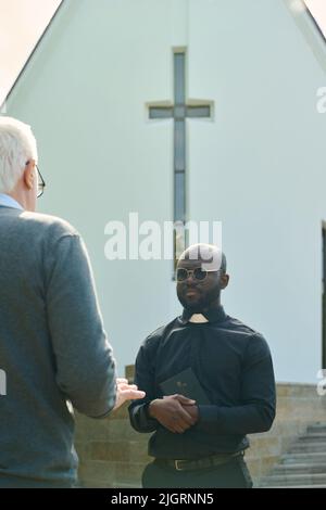Junger ernster Priester in schwarzem Hemd mit Bürokragenkragen, der die Heilige Bibel an der Brust hält und mit einem älteren männlichen Gemeindemitglied gegen das Kirchengebäude spricht Stockfoto