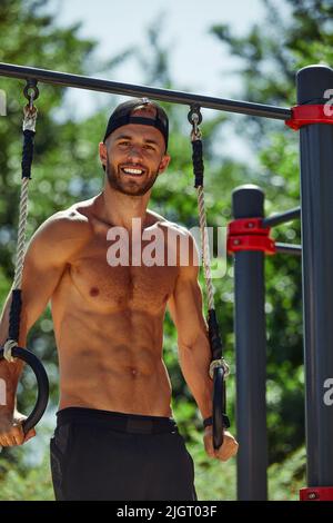 Muskulöser Sportler, der im Fitnessstudio im Freien an Gymnastikringen im Freien trainiert. Hände an Ringen tauchenden Mann, der mit Ringen trainiert Stockfoto