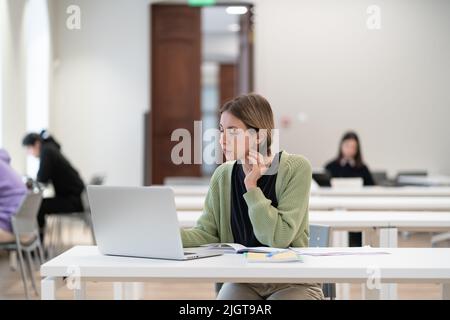 Nachdenkliche reife Studentin, die Fernlehrgänge besucht, sitzt an der Bücherei Stockfoto