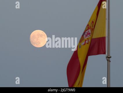 Malaga, Spanien. 12.. Juli 2022. Der Vollmond erhebt sich über dem Himmel, während eine spanische Flagge winkt. Der zweite Supermond, bekannt als „Hirscher-Supermond“, wird in der Nacht vom 12. Auf den 13. Juli zu sehen sein. Der Supermond wird aufgrund seiner Nähe zur Erde der größte Supermond des Jahres sein. Kredit: SOPA Images Limited/Alamy Live Nachrichten Stockfoto