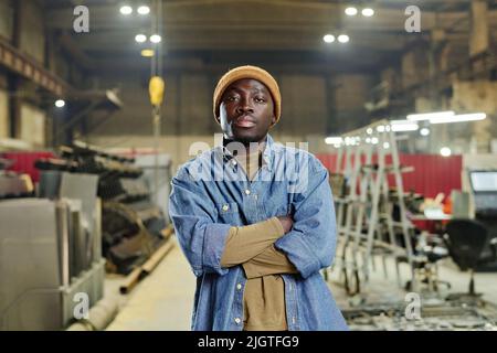 Porträt eines afrikanischen jungen Arbeiters in legerer Kleidung, der mit gekreuzten Armen in der Fabrik steht und die Kamera anschaut Stockfoto