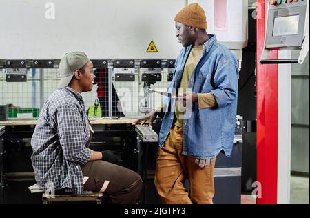 Afrikanischer junger Vorarbeiter mit digitalem Tablet, der die Arbeit einer Arbeiterin gibt, während sie an der Maschine sitzt Stockfoto