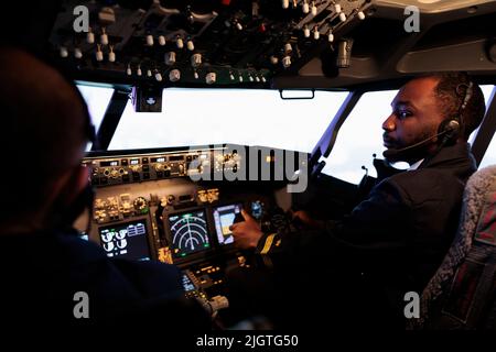 afroamerikanischer Pilot, der einen Flugzeugjet fliegt, der Teamarbeit mit dem Kapitän leistet, indem er Dashboard-Befehle und Navigation verwendet. Ein Team von Flugzeugen drückt die Tasten des Bedienfelds und den Hebel, um abzuheben und zu fliegen. Stockfoto