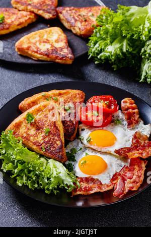 irisches Frühstück mit Spiegeleiern, Rascher- oder Speckscheiben, gebratenen Kartoffelkuchen, gegrillten Tomaten und frischem Salat auf schwarzem Teller auf Betontisch, vertic Stockfoto