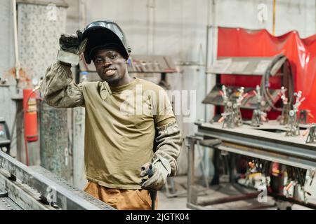 Porträt eines jungen Schweißers aus Afrika in Maske, der vor der Kamera lächelt, während er in der Fabrik mit Metallprodukten arbeitet Stockfoto