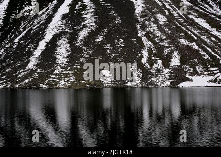 Schneestreifen, die auf schroffen schwarzen Geröll zum Schottensee abfallen, spiegeln sich an einem grauen Julitag im Kanton Graubünden in der Schweiz im kalten, dunklen Wasser des Sees wider. Der Schottensee ist ein ruhiger Rastplatz auf der Straße der Rhätischen Alpen, die Davos im Landwassertal über den Fluelapass auf 2.383 m oder 7.818 ft mit Susch im Unterengadin verbindet. Stockfoto
