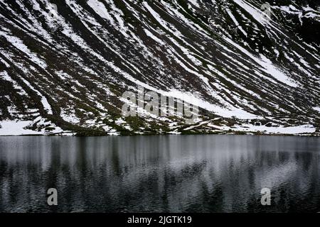 Schräge Schneestreifen, die auf schroffen schwarzen Geröll liegen, spiegeln sich an einem bewölkten Julitag im Kanton Graubünden in der Schweiz im dunklen, eisigen Wasser des Schottensees wider. Der Schottensee ist ein ruhiger Rastplatz auf der Straße der Rhätischen Alpen, die Davos über den Fluelapass auf 2.383 m Höhe mit Susch 7.818 verbindet. Stockfoto