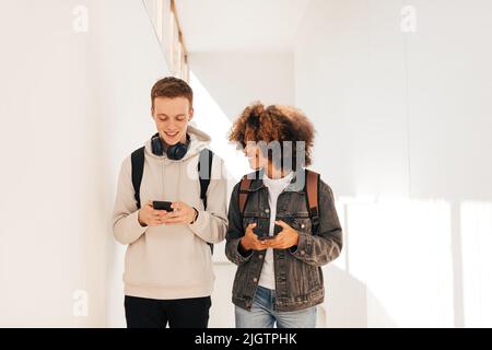Zwei lächelnde Studenten gehen im Flur und plaudern, während sie Smartphones halten Stockfoto