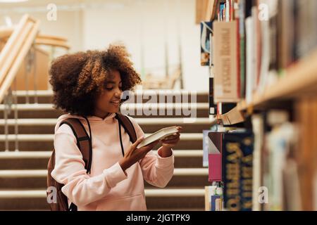 Lächelndes Mädchen mit lockigen Haaren hält ein Buch in der Nähe des Bücherregals in der Schulbibliothek Stockfoto