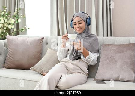 Elegante junge asiatische muslimische Frau, die einen Hijab und kabellose Kopfhörer trägt, während sie ihren morgendlichen Kaffee im Wohnzimmer schlürft. Stockfoto