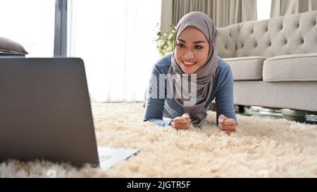 Aktive junge asiatische muslimische Frau mit Hijab, die im Wohnzimmer Übungen macht und über einen Laptop im Internet Planken trainiert. Stockfoto