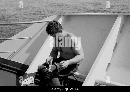 Das Foto wurde ungefähr 1961-09-22 aufgenommen. Ein Mann sitzt auf einem Schiff. Er spaltet ein Zugholz. African travel35 Bilder in Serie. Stockfoto