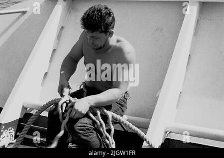 Das Foto wurde ungefähr 1961-09-22 aufgenommen. Ein Mann sitzt auf einem Schiff. Er spaltet Zugholz zusammen. African travel35 Bilder in Serie. Stockfoto