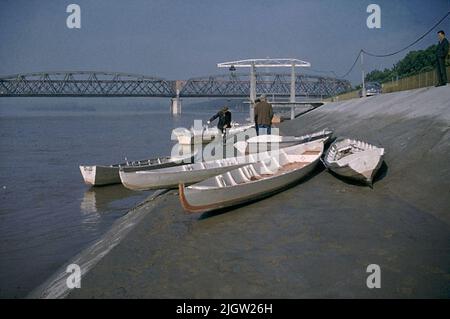 Mehrere Boote werden auf einem betonierten 'Strand' in Betrieb genommen. Drei Männer ziehen ein weiteres Boot hoch. Nach rechts geht eine Straße. Auf der Straße steht ein Auto (O. Hasslöfs Amason). Weiter weg führt eine Brücke über die river.italia (?) Stockfoto