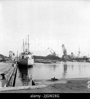 Mehrere kleine Boote befinden sich in einem Hafen. An einem Kai liegt ein Schiff. Auf dem Pier im Hintergrund sind ein paar Kraniche. Auf dem Kai auf der linken Seite sind Eisenbahnwaggons. Stockfoto