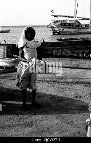 African trip, Port Sudden Acquisition: Bücher und Archivmaterial.36 Bilder in Serien. Das Foto wurde ungefähr 1961-08-22 aufgenommen. Ein junger Mann steht an einem Strand und hält zwei Muscheln. Draußen in der Bucht ist ein Schiff zum Ankern. Stockfoto