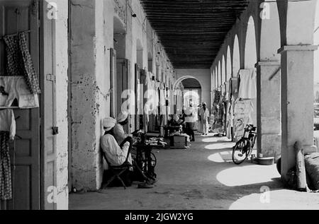African trip, Port Sudden Acquisition: Bücher und Archivmaterial.36 Bilder in Serien. Das Foto wurde ungefähr 1961-08-22 aufgenommen. Ein paar Männer sitzen in einer Arkade auf einer Geschäftsstraße. Rechts steht ein Fahrrad, das sich an einer der Säulen in der Arkade lehnt. Stockfoto