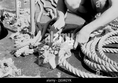 Das Foto wurde ungefähr 1961-09-22 aufgenommen. Ein Mann sitzt auf einem Schiff. Er hat Zugholz gespalten. Jetzt schneidet er die Fülle des Zuges mit einer Axt weg. African travel35 Bilder in Serie. Stockfoto