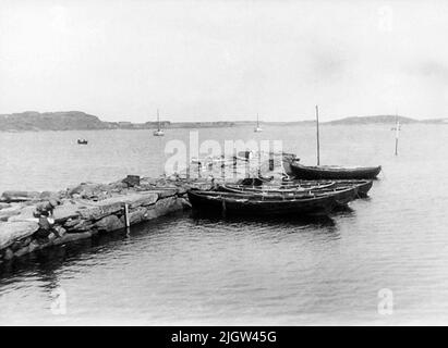 Auf der Rückseite geschrieben: Halland, Viske HD, Värö sn., Boatfjord. Kleine Boote an der Steinbrücke. Ein paar Boote liegen an einem Pier in einem Hafen. Zwei Jungen spielen auf dem Pier. Draußen in Havsviken gibt es mehrere Boote. Stockfoto