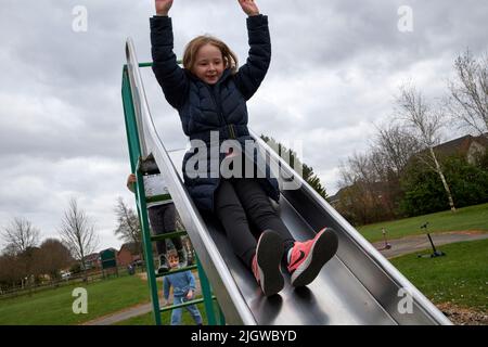 Ein junges Mädchen, das in Winterkleidung in einem Park in großbritannien auf einer Rutsche herunterrutscht und die Arme hochgezogen hat Stockfoto