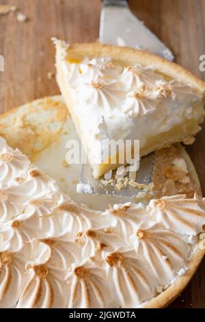Zitronenmeringue in Scheiben geschnitten und mit einem Kuchenschneider serviert. Auf einem hölzernen Schneidebrett Stockfoto