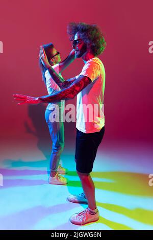 Porträt von jungen stilvollen Mann und Frau tanzen, posiert isoliert auf rotem Hintergrund in Neonlicht. Denken Sie an Tänzer Stockfoto