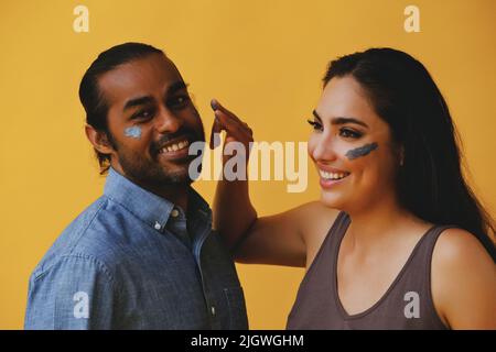 Porträt eines verspielten Paares aus der lateinamerikanischen Latina und eines indischen Mannes mit einem Tonschlamm, das mit Fingern und Händen lachend vor einem gelben Hintergrund auf das Kamerastudio blickt Stockfoto