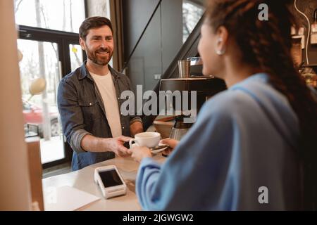Eine Barista serviert Kaffee an der Bar und gibt einem männlichen Gast eine Tasse. Cafeteria arbeiten. Stockfoto