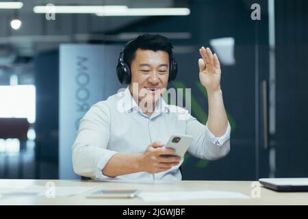 Geschäftsmann im Büro, der sich während der Arbeitspause ausruhte, Musik in großen schwarzen Kopfhörern hörte, asiatischer Mann, der Musik-App auf dem Telefon verwendete Stockfoto