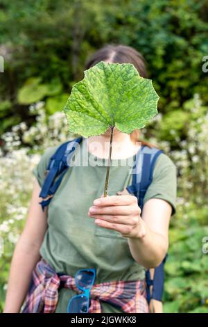 Junge Frau, die ein grünes Klettenblatt in der Hand vor ihrem Gesicht in einem gesichtslosen, anonymen psychologischen Konzeptmockup hält Stockfoto